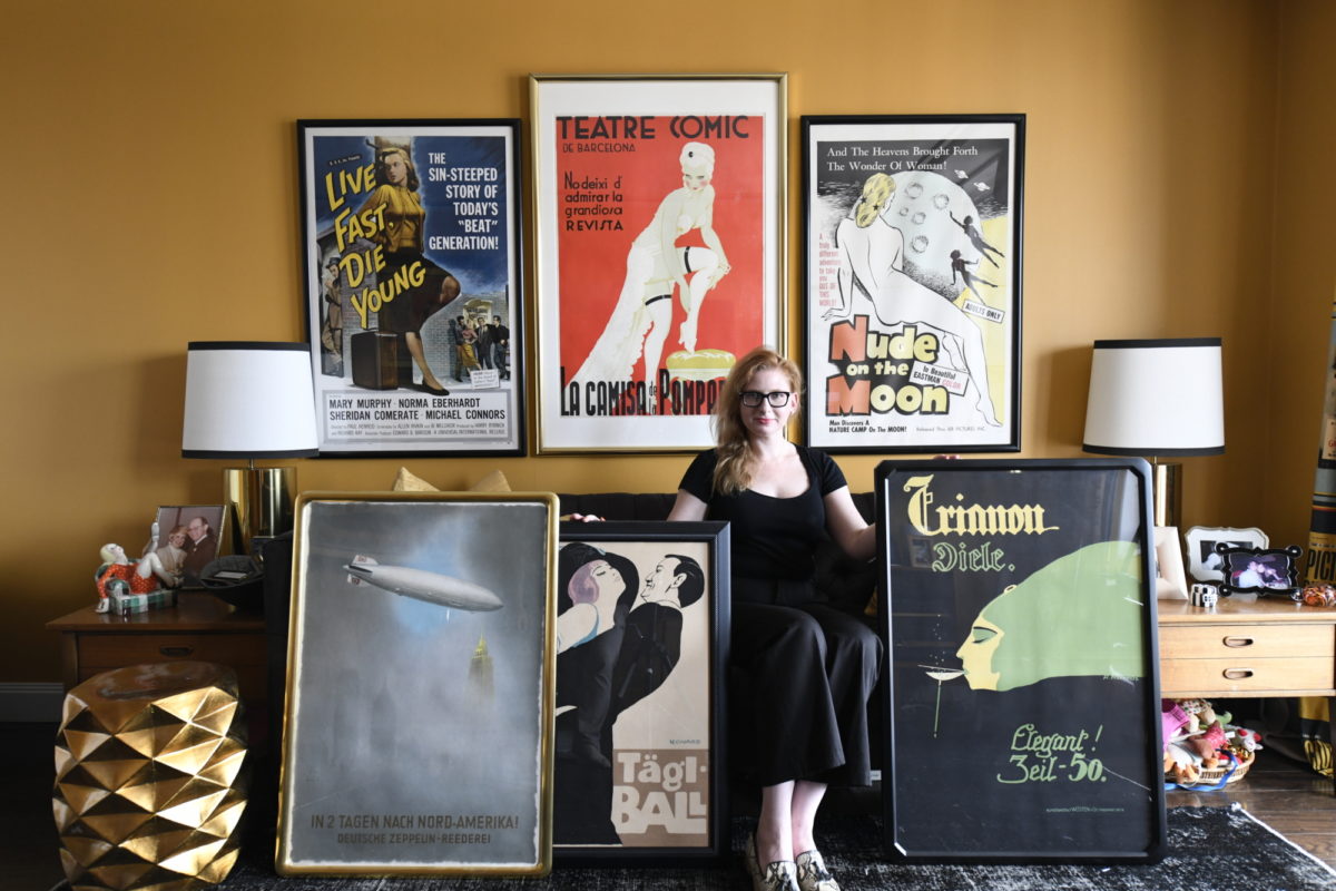 A woman is sitting on a couch while she holds 3 large posters and 3 more posters are hanging on the wall.