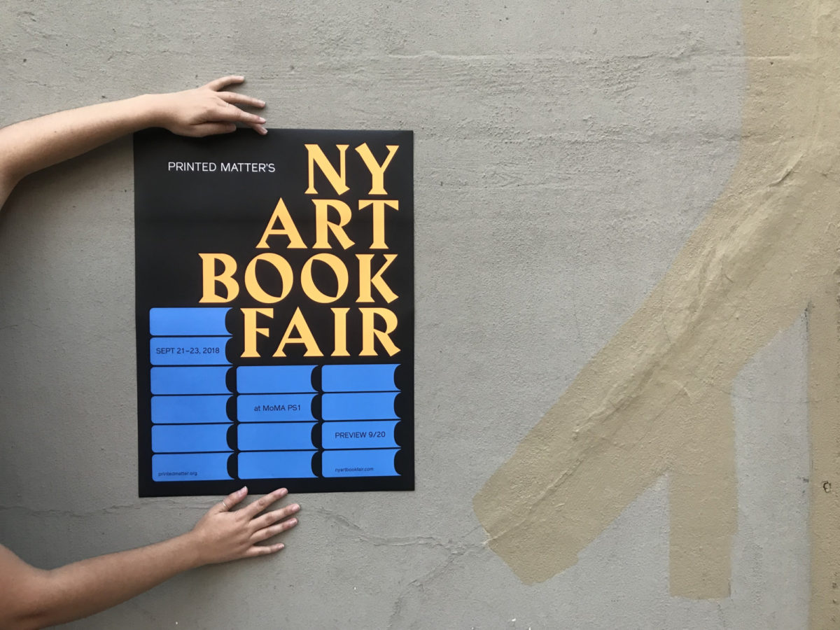 A pair of hands pasting a poster advertising the NY art book fair onto a wall.