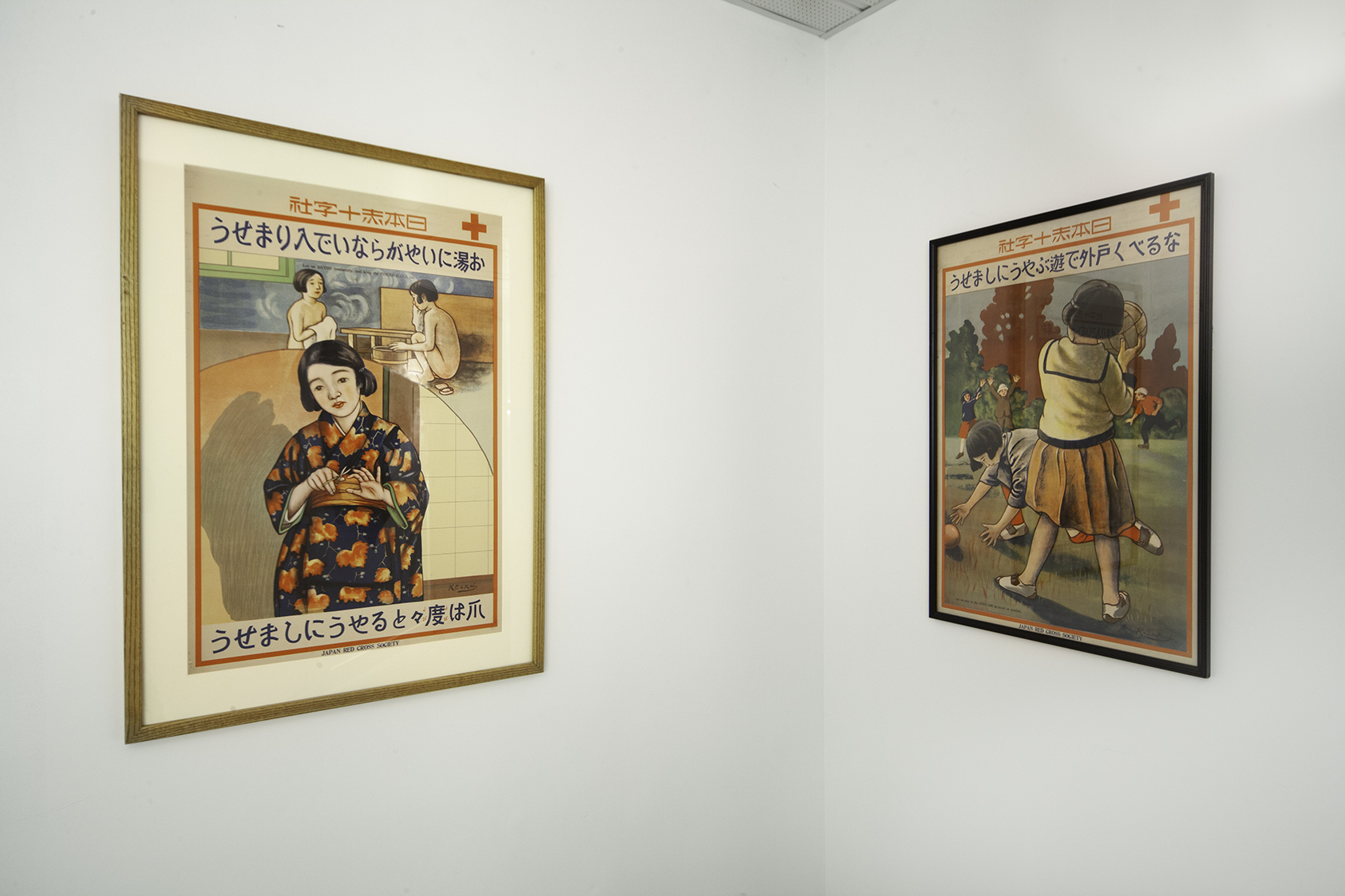 Two framed posters with Japanese script hanging in the corner of a white room. One with a young woman trimming her nails in a kimono. The other features school children playing catch in a park.