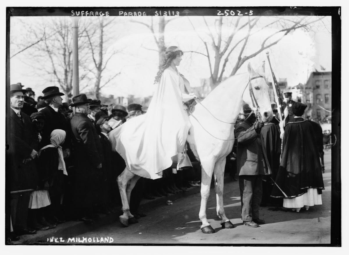black and white photograph of a woman dressed in white riding a white horse next to a crowd dressed in black