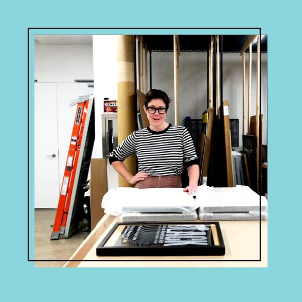 The Collections Manager smiling in front of stacked covered art frames in the archives room on a teal background.