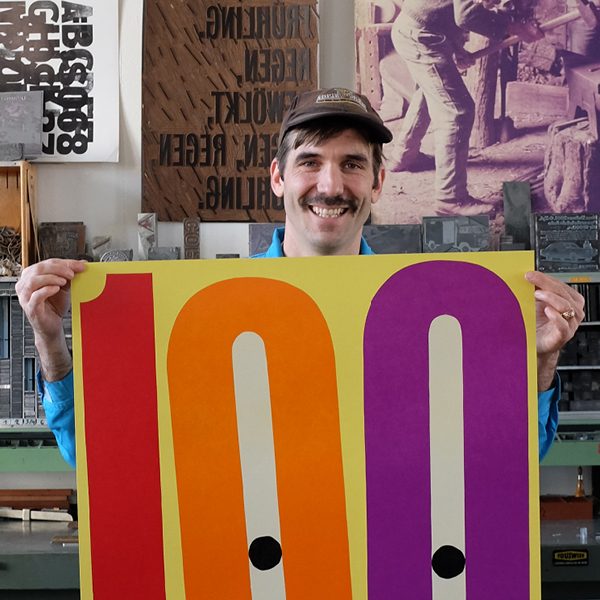 A man holding up a colorful large letterpress poster in front of his working studio.