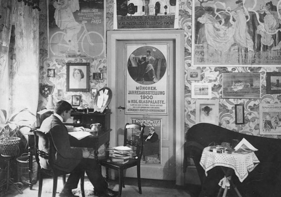 Photograph of a man sitting at a desk with his back to the viewer surrounded by posters