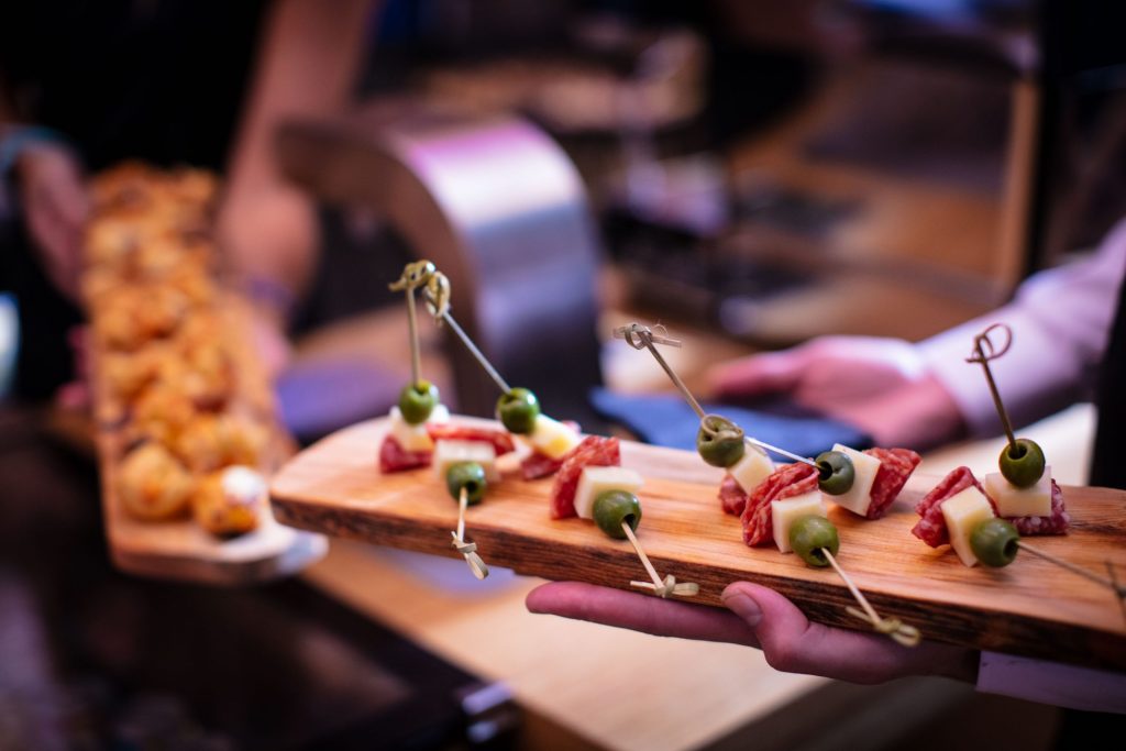 Close up of a waiter's hands presenting a wooden platter of green olive, cheese, and salami appetizers speared on toothpicks