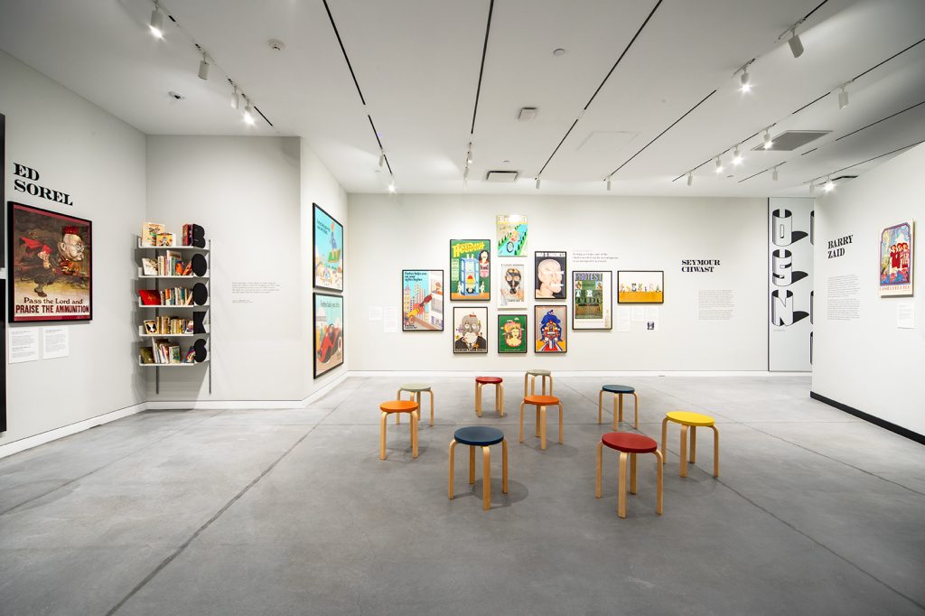 A view of a circle of colorful stools in an open gallery with white walls and colorful posters.