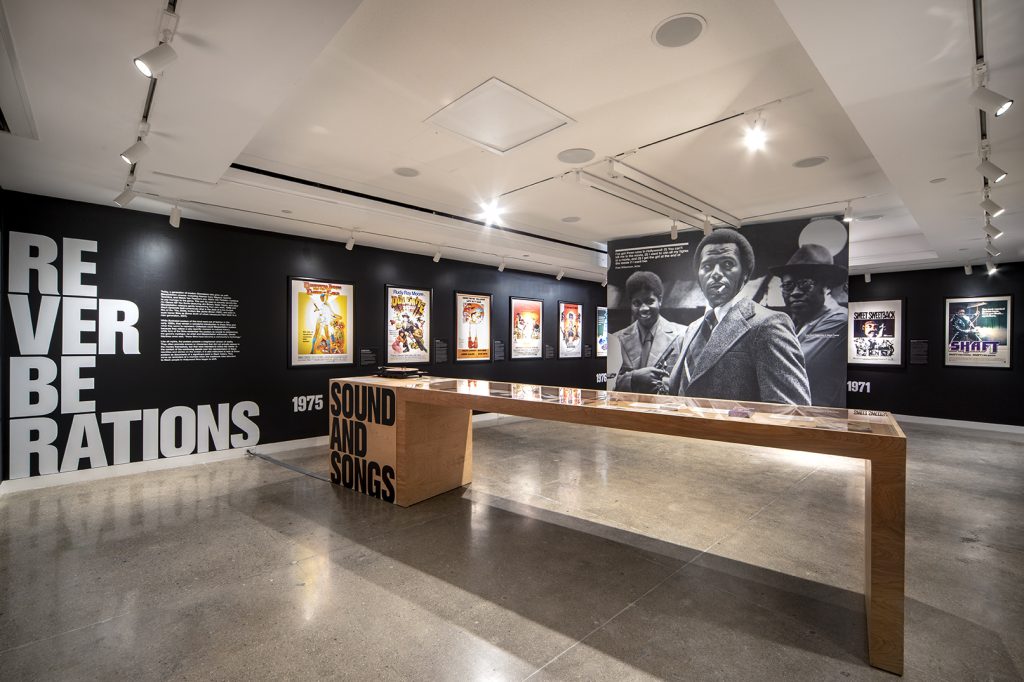 Movie posters hanging on black walls behind a long wooden display case and a large photo of Black men in suits