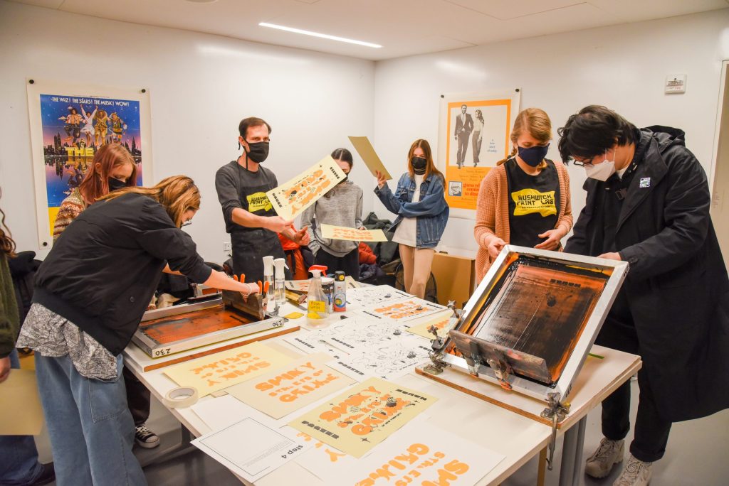 Participants taking part in a silkscreen printing workshop. Posters in various stages of completion are lying on the table.