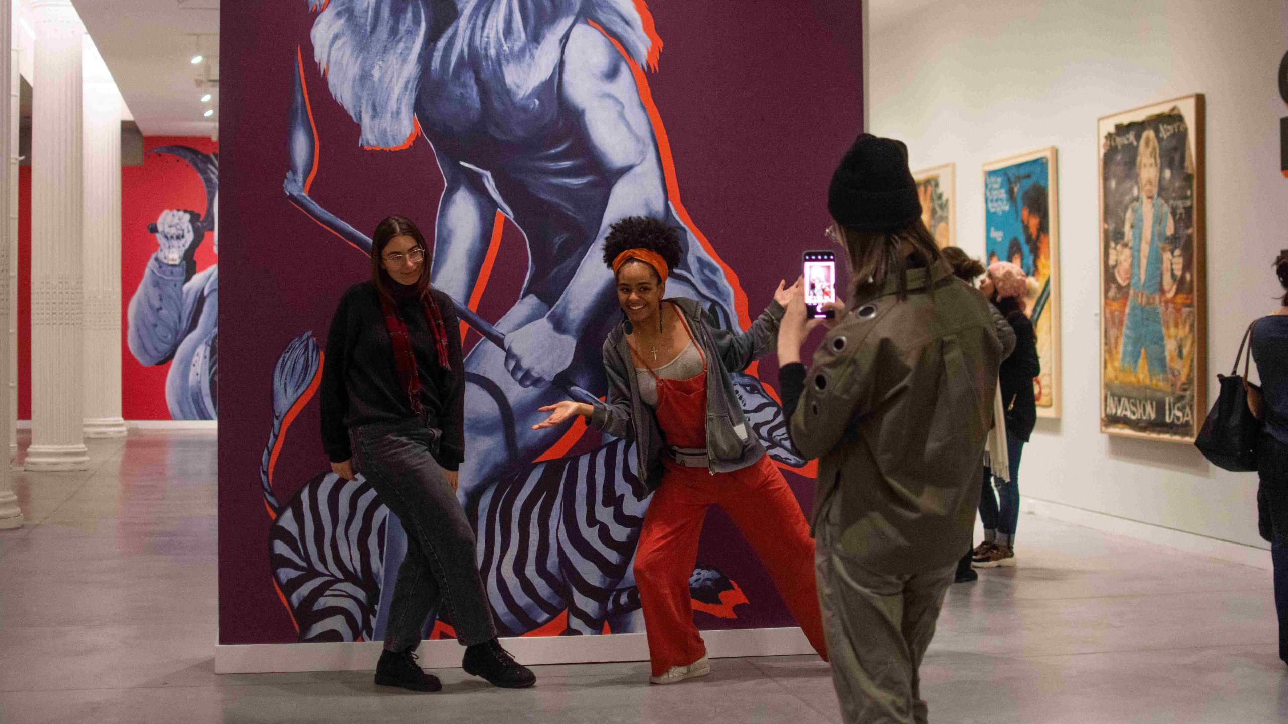 Two college students posing in a gallery while another student takes a photo of them.