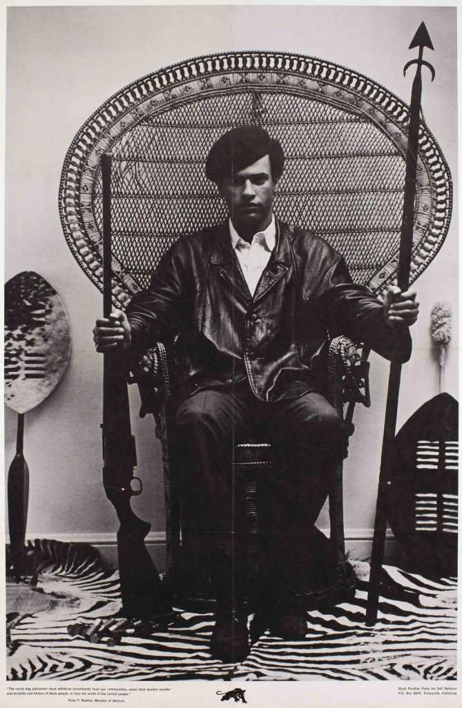 An offset poster of a black man in a peacock chair holding a spear and gun.