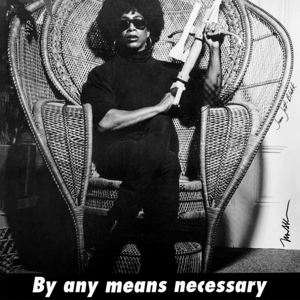 A black and white poster of a Black drag queen with a gun seated in a wicker cane chair.