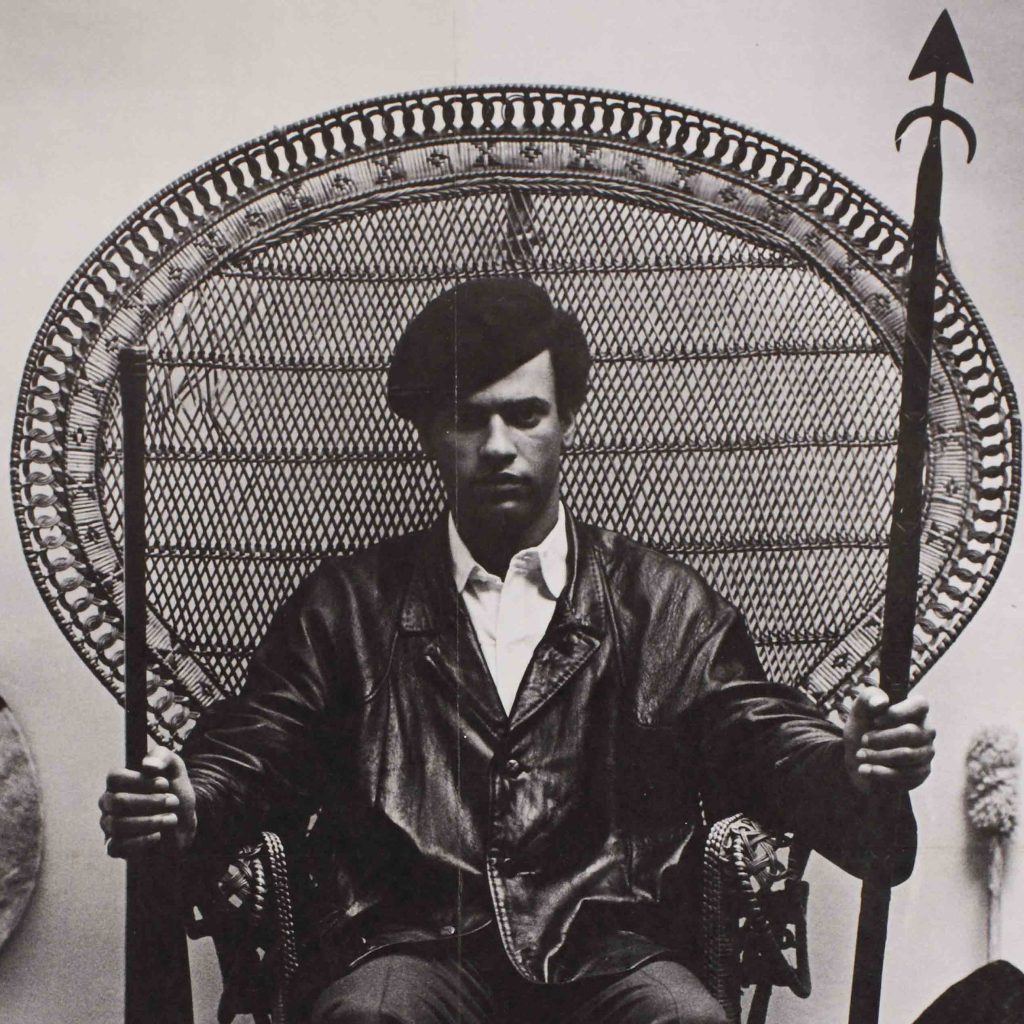 A photo of a Black man sitting in a chair holding a gun in one and a spear in the other.