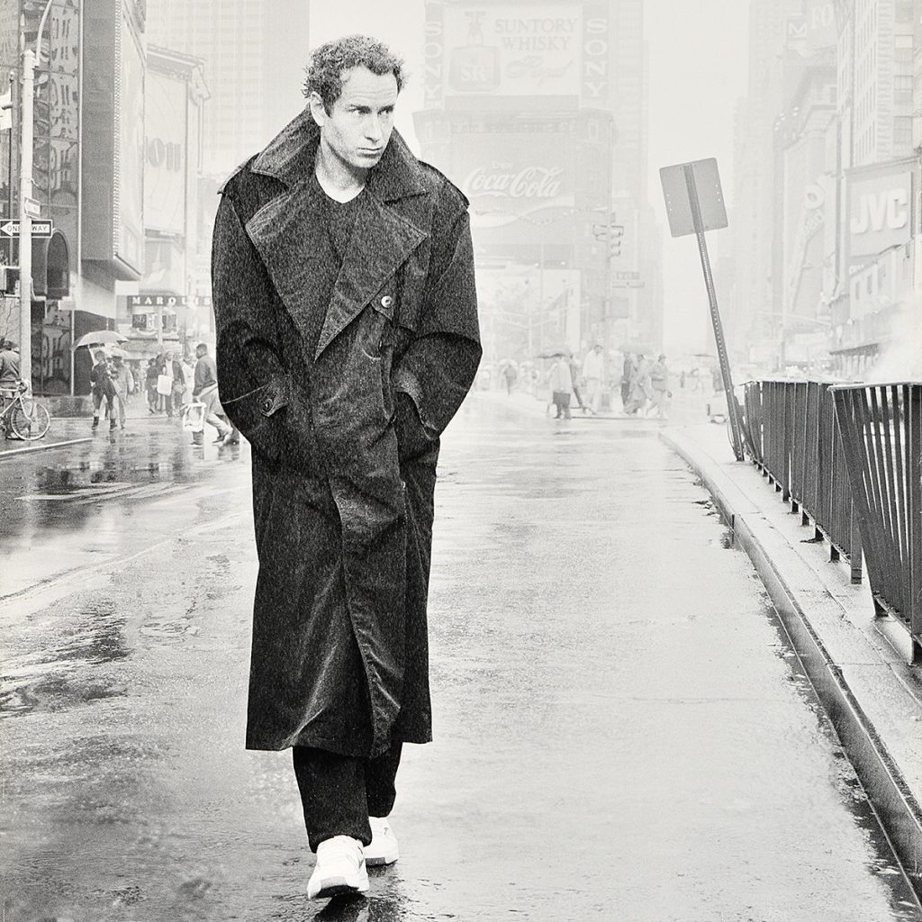 A man in a dark coat walks a street