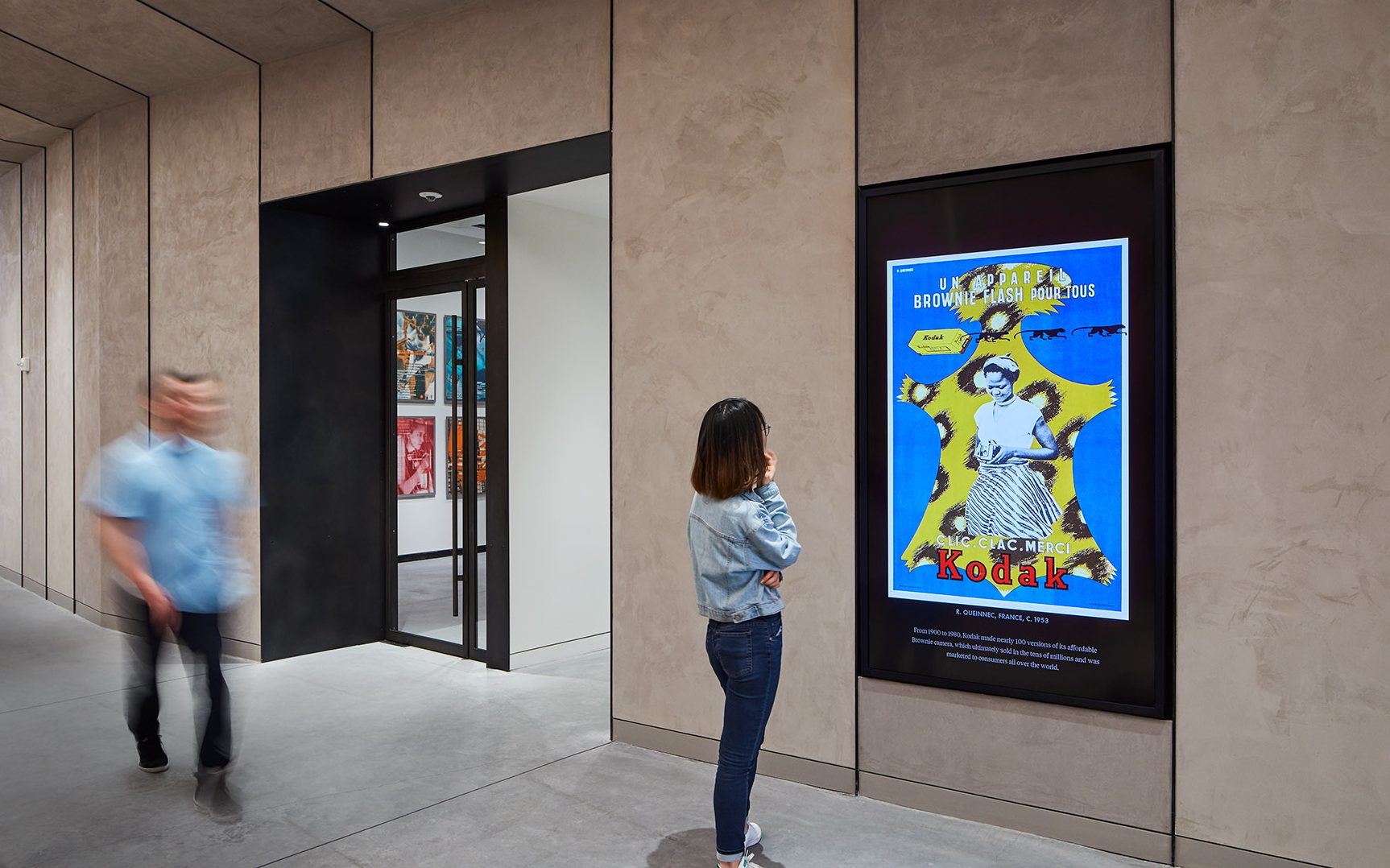 A person standing in front of the Digital Poster Wall in the Poster House foyer while another person walks by