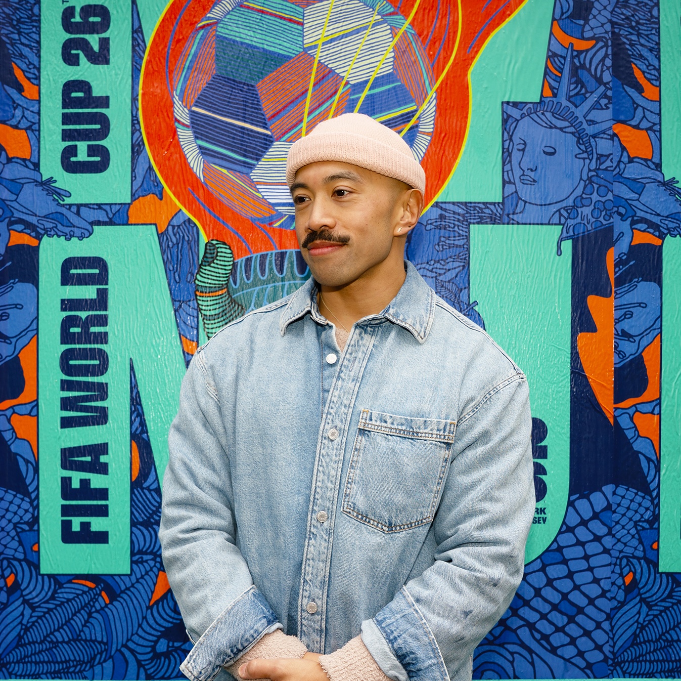 headshot of a young man wearing a beanie and a denim shirt, standing in front of a colorful poster advertising the 2026 FIFA World Cup