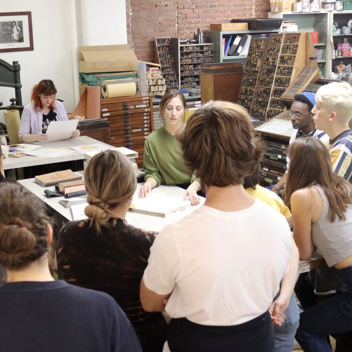 a woman teaching a bookbinding class to a group of people standing around a table