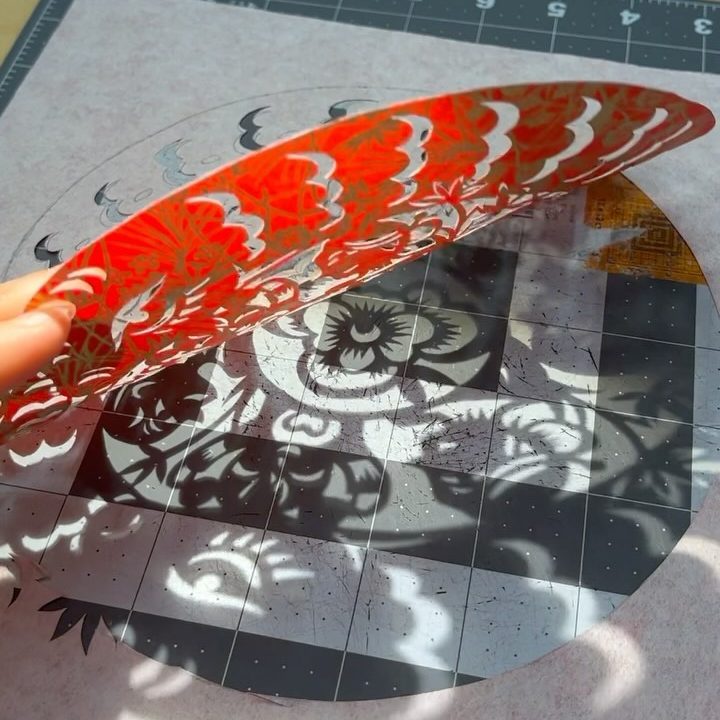a shadow of a floral motif cast on a cutting mat by an intricately cut red piece of paper being held up to the sun