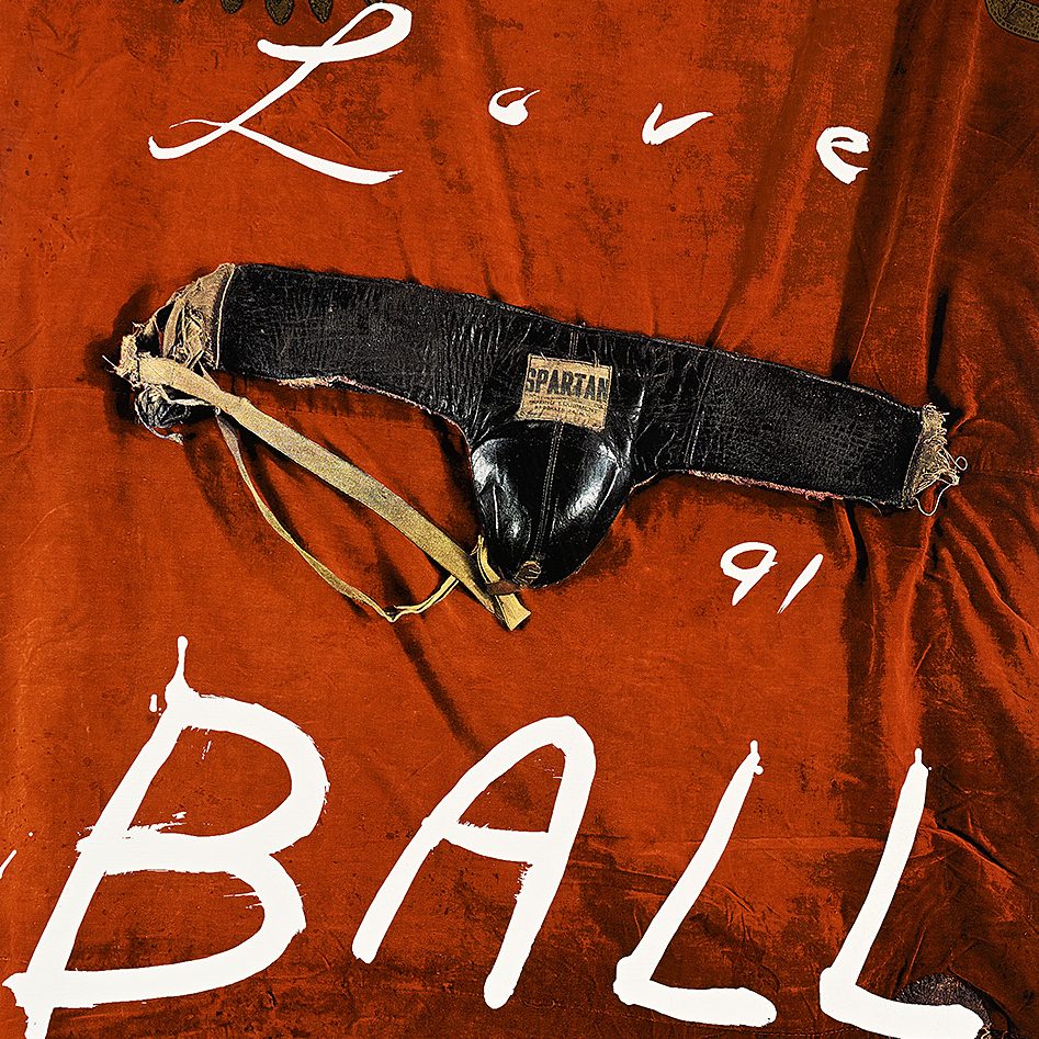 A poster of a leather jockstrap placed on a vibrant red velvet backdrop, surrounded by white handwritten text that says Love Ball.