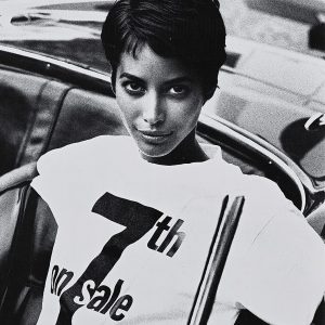 a black and white photograph of a woman with a pixie haircut wearing a white tee shirt that says 7th on Sale in black block letters