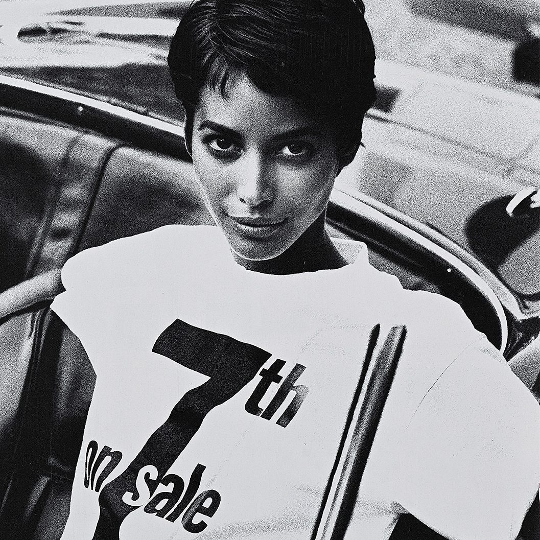 a black and white photograph of a woman with a pixie haircut wearing a white tee shirt that says 7th on Sale in black block letters