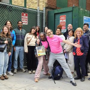 A group of smiling people standing in front of a New York City green shed.