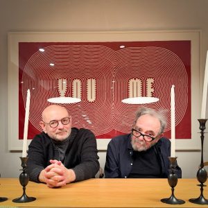 Two men sitting at a table in front of a framed print that says the words "me" and "you".