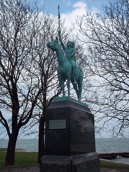 An image of a bronze sculpture in a faded blue-green color of an indigenous man on a horse holding a staff above him, in a park with trees and a blue sky